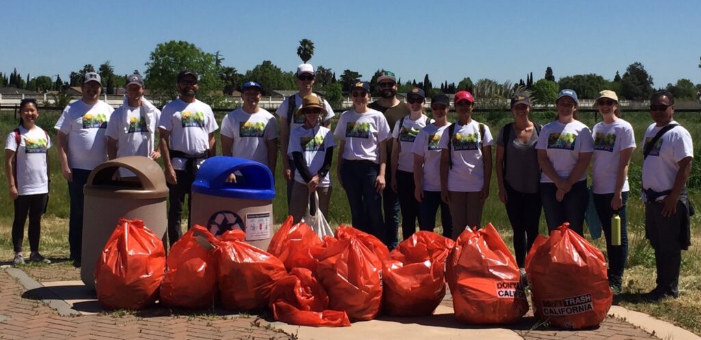 A group of 16 people standing outside after the Creek Week Waterway Cleanup. They are shown with two trash cans and 10 large orange trash bags that say "Don't Trash California."