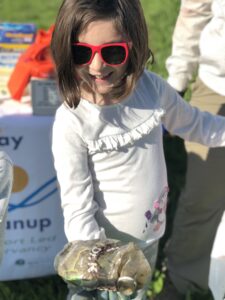 A young girl wearing red sunglasses and blue gloves holding up an old plastic soda bottle toward the camera during a Waterway Cleanup.