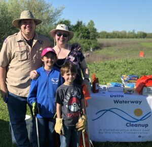 A family with an adult man, adult woman, and two young boys who are wearing gloves and holding trash grabbers outside next to the "Delta Waterway Cleanup" banner.