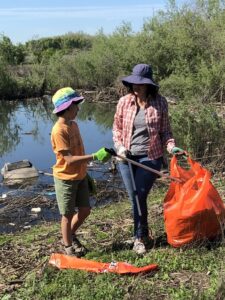 An adult woman and a young boy near a creek using trash grabbers to place trash into a large orange plastic trash bag.