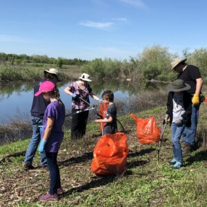 Six people, adults and children, standing outside next to a creek using trash grabbers to place trash into large orange plastic trash bags.