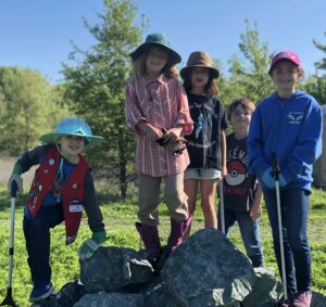 Five children, some wearing gloves and some holding trash grabbers, smiling at the camera while standing on a pile of grey rocks in the middle of the green grass at the Waterway Cleanup.